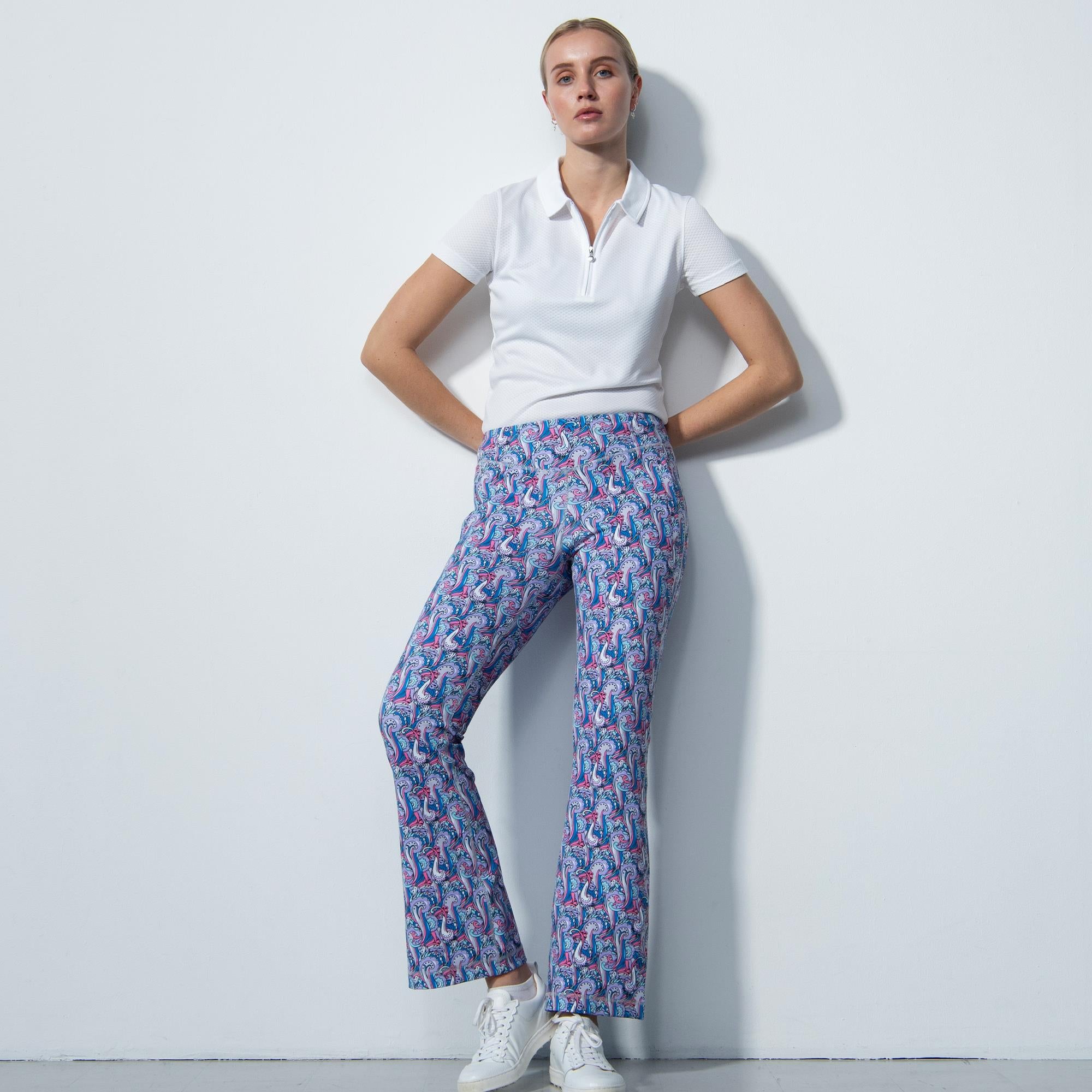 Studio shot of blonde woman modelling Daily Sports patterned flared trousers in purple, pink and blue, with a white plain polo shirt  